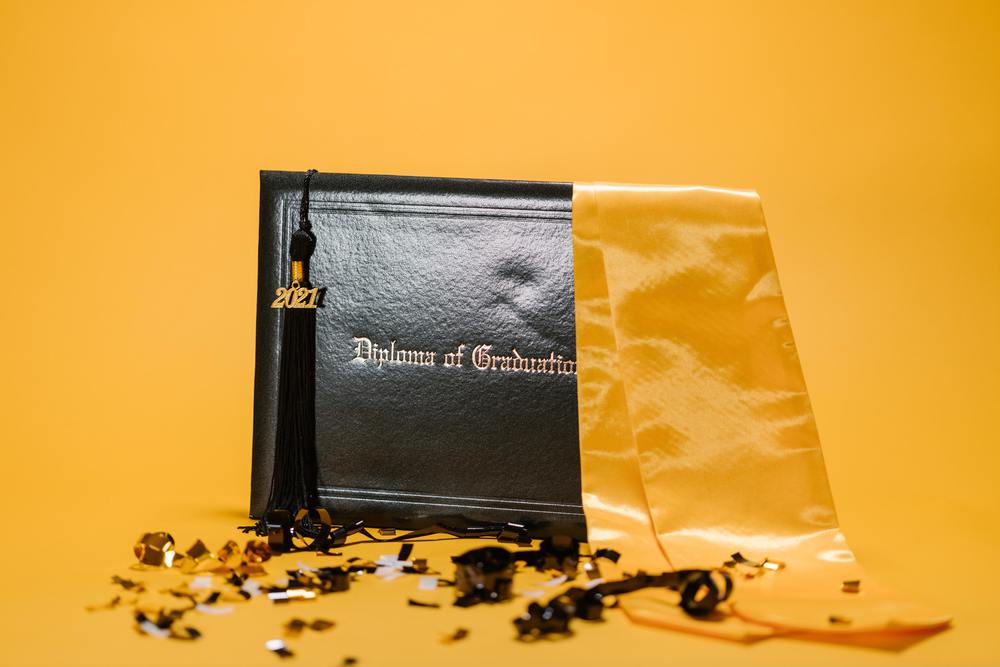 An image of a diploma, tassel and sash in gold, on a gold backdrop surrounded by confetti